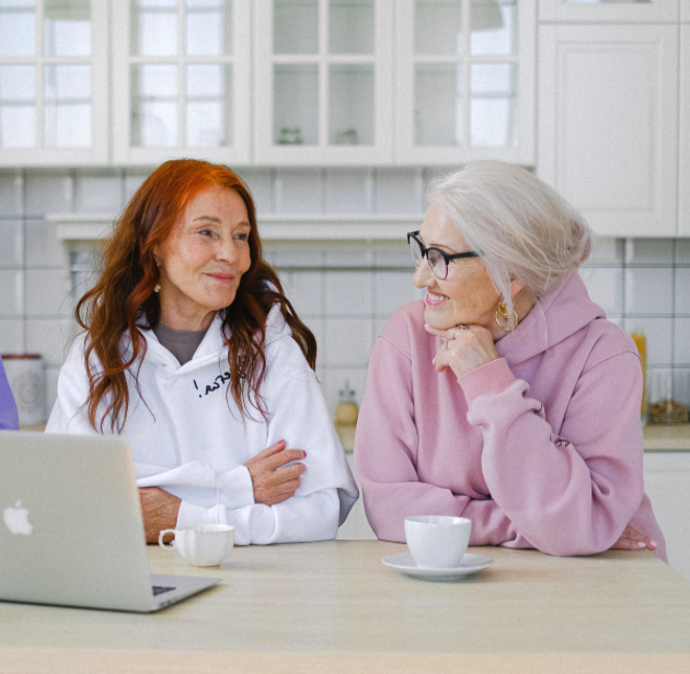 Two elderly woman smiling at each other in a kitchen in front of a laptop